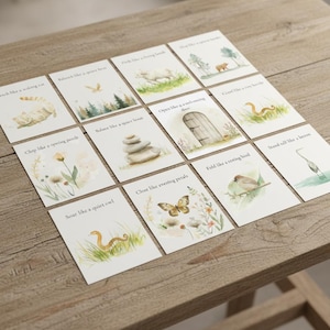 May include: Twelve small cards with watercolour illustrations and text are arranged on a wooden surface. Each card displays a different image, such as a cat, a bird, a snake, and a butterfly, with phrases like "Stretch like a waking cat" and "Soar like a quiet owl."