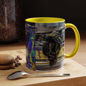 Peut inclure: Une tasse à café à poignée jaune avec un design saisissant. Un côté présente un portrait coloré, l'autre une moto en action. L'intérieur de la tasse est jaune, posée sur une surface en bois.