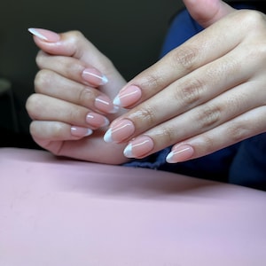 May include: Oval-shaped nails with a classic French manicure design. The nails have a natural pink base with white tips. The hands are positioned on a light pink surface.