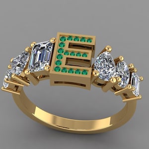 Peut inclure: Une bague en or avec la lettre "E" ornée de petites pierres vertes. La bague est ornée de plusieurs pierres précieuses claires et étincelantes de différentes formes, serties dans un anneau en or. Fond neutre.