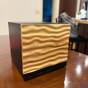 May include: A square electronic device featuring a matte black housing and a light-colored wooden front panel with a horizontal, wavy, three-dimensional texture. The base of the black unit includes visible ports along the bottom edge.