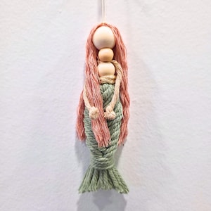May include: A handmade macrame mermaid doll with a wooden head and flowing pink hair. The mermaid's body is crafted from green macrame rope, with a detailed tail. The doll is suspended by a white string, perfect for hanging as a decorative item.