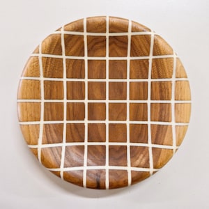 Può includere: Piatto rotondo in legno con motivo a griglia. Il piatto è realizzato in legno marrone chiaro e presenta un design a griglia bianca sulla sua superficie. Un oggetto decorativo.