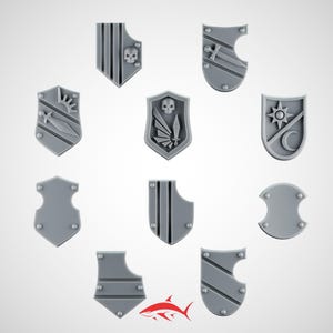 May include: A collection of nine gray miniature shields with various designs. Some shields feature stripes, skulls, swords, suns, and moons. Each shield has small, round rivets. The shields are arranged on a white background.