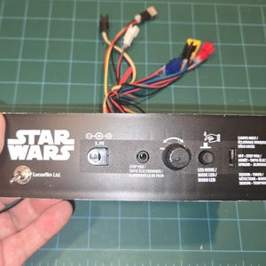 May include: A black Star Wars electronic device with various ports, a dial, and a switch. The device has the Star Wars logo and text. Wires with colored connectors extend from the top. An antenna is attached.