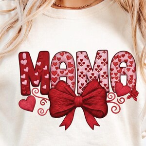 Può includere: T-shirt color crema con la scritta "MAMA" in lettere rosse riempite di motivi a cuore. Un grande fiocco rosso è centrato sotto la parola, con volute e cuori decorativi. Una rosa è sul lato destro.
