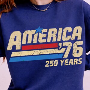 May include: Navy blue sweatshirt featuring the text "AMERICA '76 250 YEARS" in a distressed, vintage style. The design incorporates a star, and red, white, and blue stripes, celebrating the nation's history.