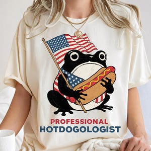 Puede incluir: Camiseta color crema con una rana de dibujos animados sosteniendo un perrito caliente y una bandera estadounidense. La rana es negra y blanca con detalles en rojo y azul. El texto "PROFESSIONAL HOTDOGOLOGIST" está impreso debajo en rojo y azul.