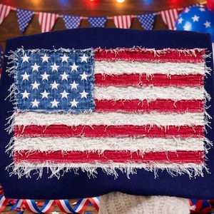 Op de afbeelding: Een marineblauw decoratief kussen met een distressed Amerikaans vlagontwerp. De vlag heeft een denimblauwe vierkant met witte sterren en rood-witte strepen. Ideaal voor patriottische woondecoratie.