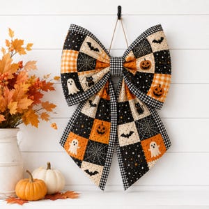 May include: A large decorative bow made of quilted fabric featuring a patchwork pattern of black, orange, and cream squares. The design includes motifs of ghosts, pumpkins, black cats, bats, and spiderwebs, all bordered by a black and white gingham trim.