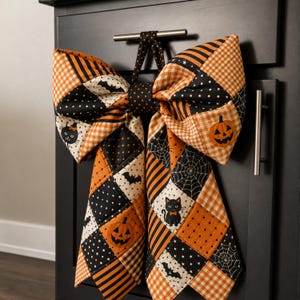 May include: A large, quilted fabric bow featuring a patchwork design in black and orange. The pattern includes motifs of jack-o'-lanterns, black cats, bats, spiderwebs, stars, polka dots, and gingham checks. The bow is suspended by a fabric loop.