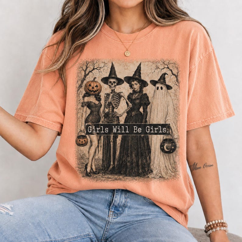 May include: A peach-colored t-shirt featuring a graphic print of four figures in witch hats: a person with a pumpkin head, a skeleton, a woman in a long dress, and a ghost. The text Girls Will Be Girls is printed in a black rectangular box.