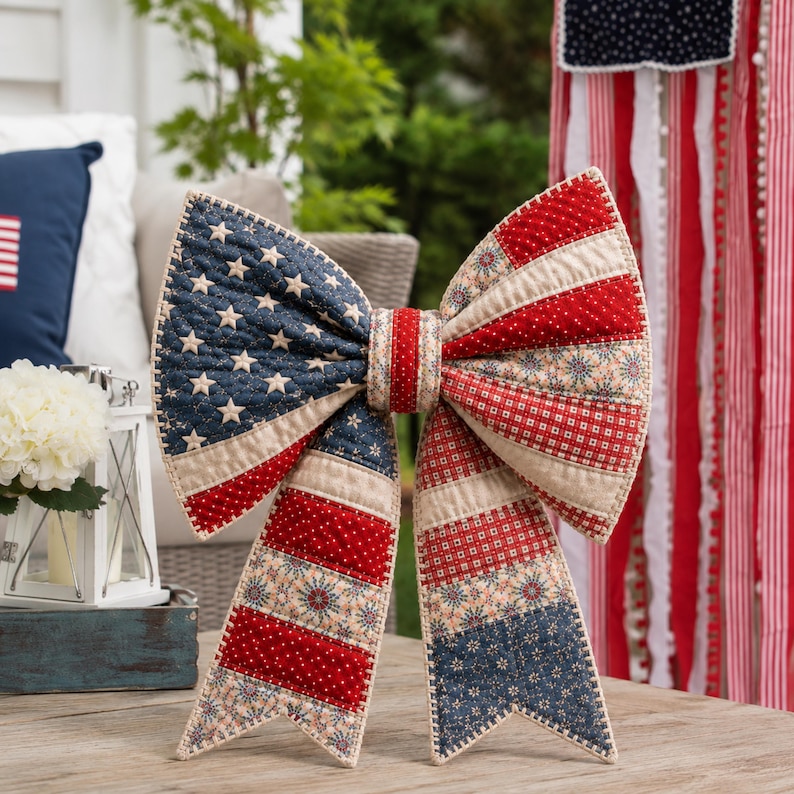 K&ouml;nnte beinhalten: Eine gro&szlig;e, dekorative Schleife im patriotischen Stil. Die Schleife hat ein Patchwork-Design mit roten, wei&szlig;en und blauen Stoffen, einschlie&szlig;lich Sternen und Streifen. Die Schleife ist aus Stoff und dient wahrscheinlich der Dekoration.