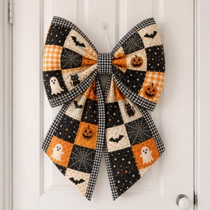May include: A large decorative bow made of quilted fabric featuring a patchwork pattern of Halloween motifs including orange pumpkins, white ghosts, black cats, bats, and spiderwebs on black, orange, and cream backgrounds with black and white gingham trim.