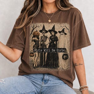 May include: A brown short-sleeve t-shirt featuring a graphic print of four figures in witch hats: a person with a pumpkin head, a skeleton, a person in a dark dress, and a ghost. The text Girls Will Be Girls is printed across the center.