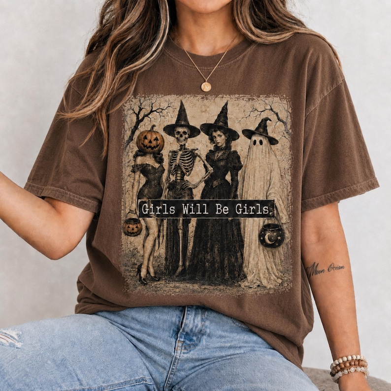 May include: A brown short-sleeve t-shirt featuring a graphic print of four figures in witch hats: a person with a pumpkin head, a skeleton, a person in a dark dress, and a ghost. The text Girls Will Be Girls is printed across the center.