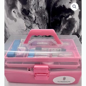 May include: A pink phlebotomy kit with a clear lid and a pink handle. The kit contains various medical supplies, including vials and pens, organized in compartments. The kit has a white oval sticker with the text "The Phlebotomy Kit".
