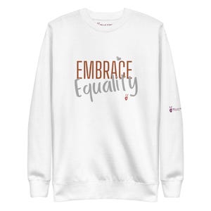 May include: A white crewneck sweatshirt featuring the text EMBRACE in brown and Equality in gray. A small gray heart sits above the text, and a small red hand symbol is below. The sleeve features a small logo with the text BELLA STRO.