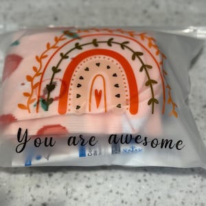 Könnte beinhalten: Eine transparente Plastiktüte mit einem Paar Socken mit Regenbogenmuster. Auf der Tüte steht der Schriftzug "You are awesome". Der Regenbogen ist orange, pfirsichfarben und weiß.