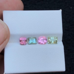 May include: Four colorful gemstones displayed in a white box. The gems include a pink, a teal, another pink, and a light green stone. The stones are rectangular and square cut. The gems are displayed against a dark background.