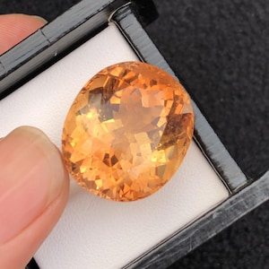 May include: An oval-shaped, faceted gemstone with a warm, orange hue. The gem is set in a black box with a white interior, and is held by a person's fingers. The gem's facets catch the light, creating a sparkling effect.