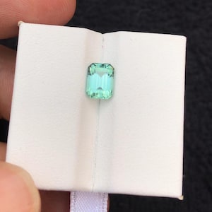 May include: A rectangular, light teal gemstone with a faceted cut. The gem is set against a white background, held by a hand. The gem's colour is a vibrant, cool teal, with visible internal facets.