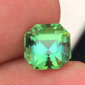 Pode incluir: Uma pedra preciosa verde esmeralda facetada com formato octogonal. A gema tem um corte brilhante, refletindo a luz e criando um efeito cintilante. A pedra é segurada entre dois dedos, mostrando seu tamanho e clareza.