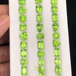 May include: A collection of vibrant, lime green peridot gemstones in various shapes, including round, pear, and emerald cuts. The gemstones are displayed on a white surface, showcasing their clarity and brilliance. The peridots are arranged in neat rows.