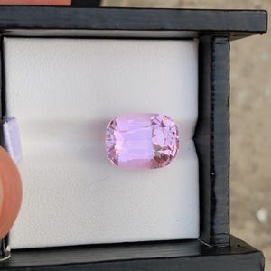 May include: A light pink, cushion-cut gemstone is displayed in a black box with a white interior. The gemstone has a rectangular shape with rounded corners and multiple facets, reflecting light. The box is open, revealing the gem.