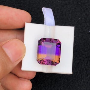 May include: A large, rectangular gemstone with a gradient of colors, including purple, orange, and pink. The gem is held in a white box with a white ribbon. The gem is faceted, reflecting light.