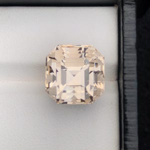 May include: A faceted, light peach-colored gemstone with an emerald cut, displayed in a black box with a white interior. The gem has a square shape with beveled edges and internal reflections.
