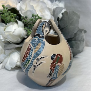 Puede incluir: Jarrón de cerámica color crema con dos asas, adornado con diseños de loros coloridos. Los loros presentan detalles azules, rojos y marrones. El jarrón tiene forma redondeada y probablemente está hecho a mano.