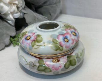 May include: A small, two-piece, white ceramic container with a lid. The container and lid are decorated with pink, blue, and yellow floral designs and green leaves. The rim of the container is trimmed with gold.