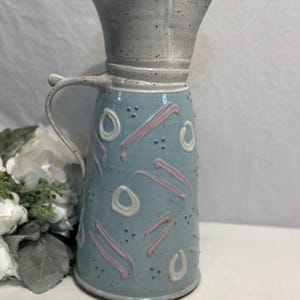 Puede incluir: Jarra de cerámica hecha a mano con cuerpo azul claro, adornada con diseños abstractos rosas y blancos. La jarra presenta un asa y una parte superior acampanada única en un acabado gris moteado. Ideal para servir bebidas.