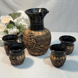 May include: A handmade ceramic sake set featuring a tall, textured pitcher with a black glazed top and four matching small cups. The set has a rustic, earthy aesthetic with brown and black tones.