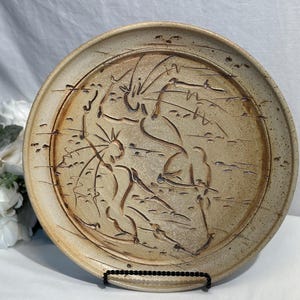 Può includere: Un piatto in ceramica beige con un disegno astratto in rilievo marrone. Il piatto ha un bordo largo ed è esposto su un supporto nero. Il design sembra essere a tema naturale.