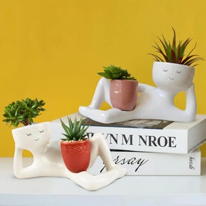May include: Two white ceramic planters shaped like reclining figures, each holding a small potted succulent. The planters have a minimalist design with closed eyes and a smiling face. One holds a red pot, the other a pink pot.