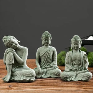 Könnte beinhalten: Drei grüne Buddha-Statuen aus Stein in verschiedenen Sitzpositionen. Eine lehnt sich an, eine andere meditiert, und die dritte hat eine erhobene Hand. Die Statuen stehen auf einer Holzoberfläche.