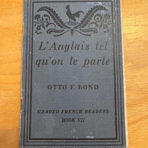 May include: A vintage book titled "L'Anglais tel qu'on le parle" by Otto F. Bond. The cover is a faded blue with black text and decorative elements, including a rooster. It is part of the "Graded French Readers" series, Book VII.