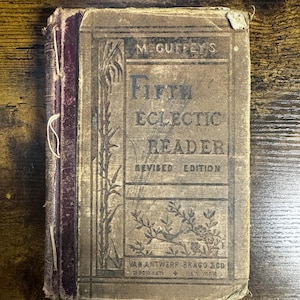 Może przedstawiać: Zniszczony, zabytkowy egzemplarz McGuffey's Fifth Eclectic Reader, Revised Edition. Okładka jest w kolorze beżowym z czarnym tekstem i ilustracjami, w tym ptakiem i kwiatowym wzorem. Grzbiet jest bordowy, a książka nosi ślady wieku i użytkowania.
