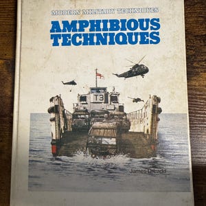 May include: A vintage book titled "Amphibious Techniques" with a cover illustration of a military landing craft and helicopters. The book is part of the "Modern Military Techniques" series, by James D. Ladd.
