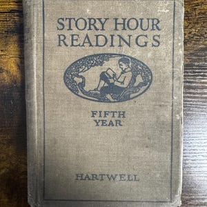 May include: A vintage book titled "Story Hour Readings" with a light brown cover. The cover features an illustration of a person reading under a tree and the text "Fifth Year" and "Hartwell". The book has a worn appearance.