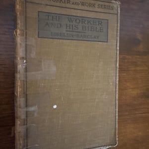 Könnte beinhalten: Ein antikes Buch mit dem Titel "The Worker and His Bible" aus der "Worker and Work Series". Der Einband ist in einem verblichenen Beigeton gehalten, der Titel in einem dunkleren Farbton. Das Buch weist Altersspuren auf, mit Klebeband am Rücken und an den Kanten.
