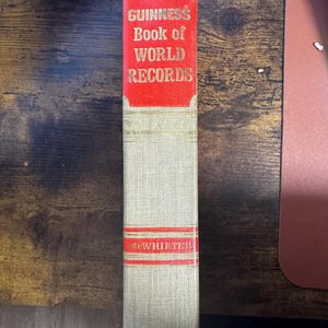 May include: A hardcover book titled "Guinness Book of World Records." The book has a red rectangular label with gold lettering. The spine is beige with red accents and the name "McWHIRTER" printed on it.