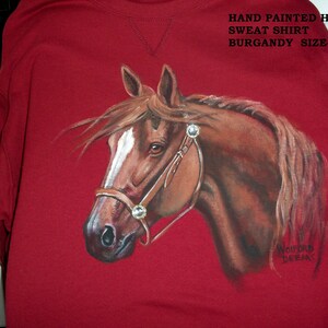 May include: Burgundy sweatshirt with a hand-painted image of a brown horse's head. The horse is wearing a brown leather bridle with silver accents. The text "HAND PAINTED HORSE SWEAT SHIRT BURGANDY SIZE-LG" is printed on the top of the sweatshirt.