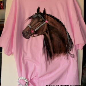 May include: A pink t-shirt with a hand-painted brown horse design. The horse has a pink bridle and pink rhinestones. The shirt is a tunic style and says "Hand Painted Horse T-Shirt One Size Fits Most Tunic Style".
