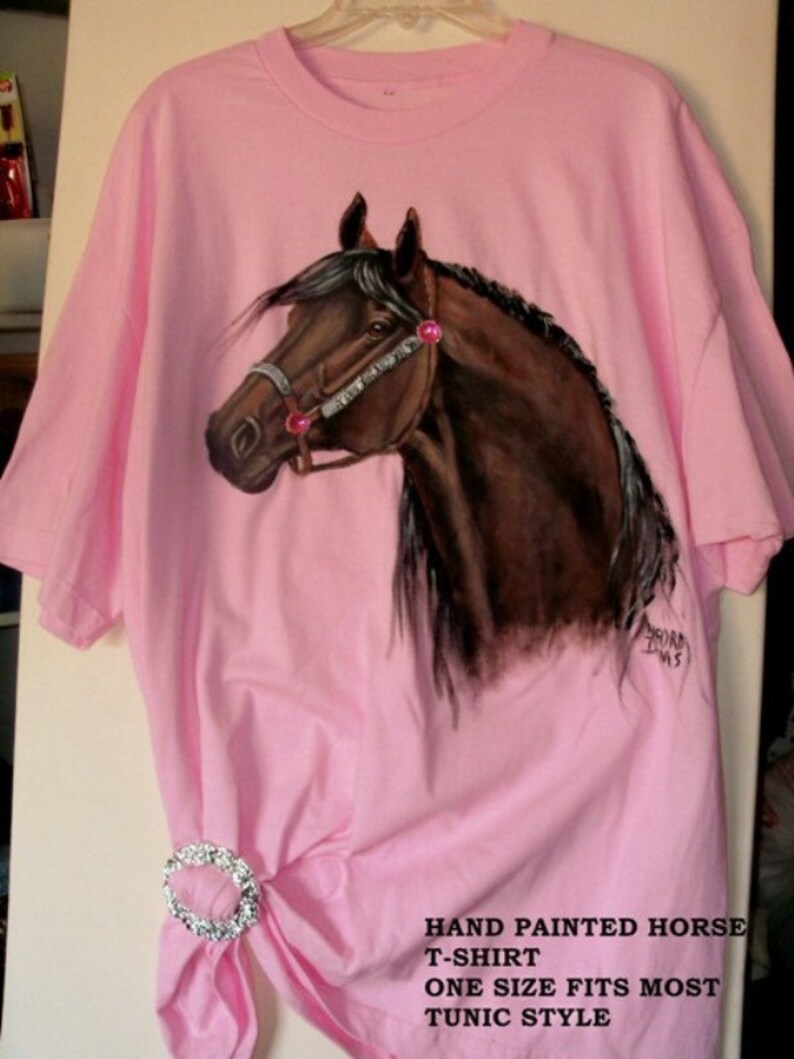 May include: A pink t-shirt with a hand-painted brown horse design. The horse has a pink bridle and pink rhinestones. The shirt is a tunic style and says "Hand Painted Horse T-Shirt One Size Fits Most Tunic Style".