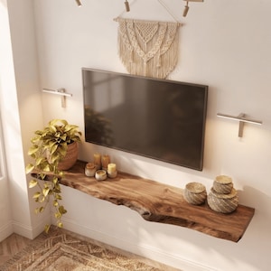 Könnte beinhalten: Ein schwebendes Wandregal aus Holz mit Naturkante unter einem Flachbildfernseher. Auf dem Regal stehen eine hängende Topfpflanze, vier kleine zylindrische Kerzen und zwei geflochtene Körbe. Über dem Fernseher befindet sich ein Makramee-Wandbehang.