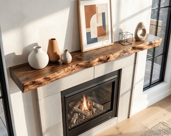 May include: A live-edge wooden fireplace mantel shelf featuring a natural wood grain finish. The mantel is decorated with a white ceramic vase, a tall tan vase, a small speckled vase, a framed abstract print, a black wire cube, and a stone sculpture.