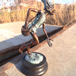 Op de afbeelding: Een bronzen sculptuur van een grillige elfenfiguur, met een buidel. De elf wordt in beweging afgebeeld, met een puntmuts en gedetailleerde gelaatstrekken. De sculptuur is gemonteerd op een ronde, donkere basis en heeft een patina.